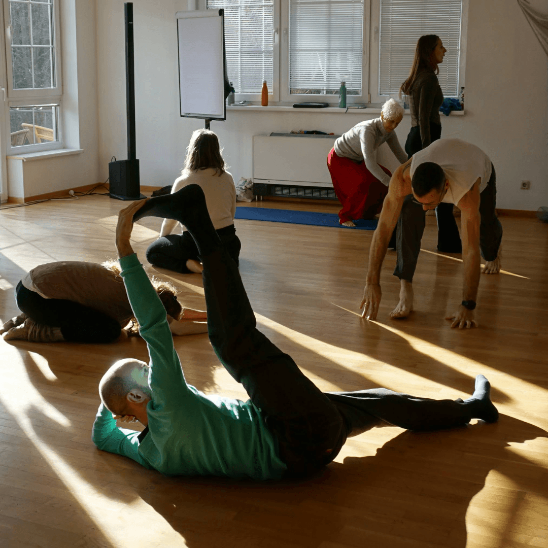 People moving on wooden dance floor, person lying down hand touching toes of leg stretched up.